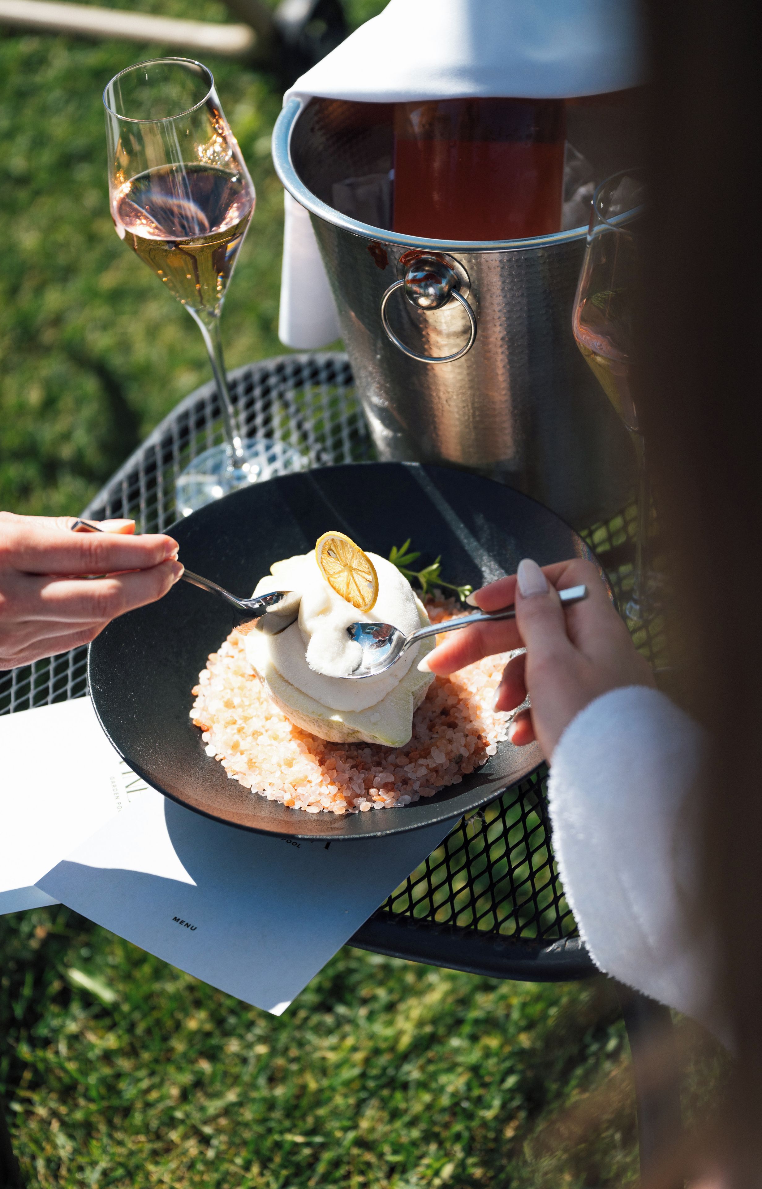 Una persona corta un postre con un tenedor sobre un plato negro, con una copa de vino blanco y un recipiente metálico al fondo.