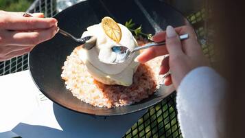 Una persona corta un postre con un tenedor sobre un plato negro, con una copa de vino blanco y un recipiente metálico al fondo.
