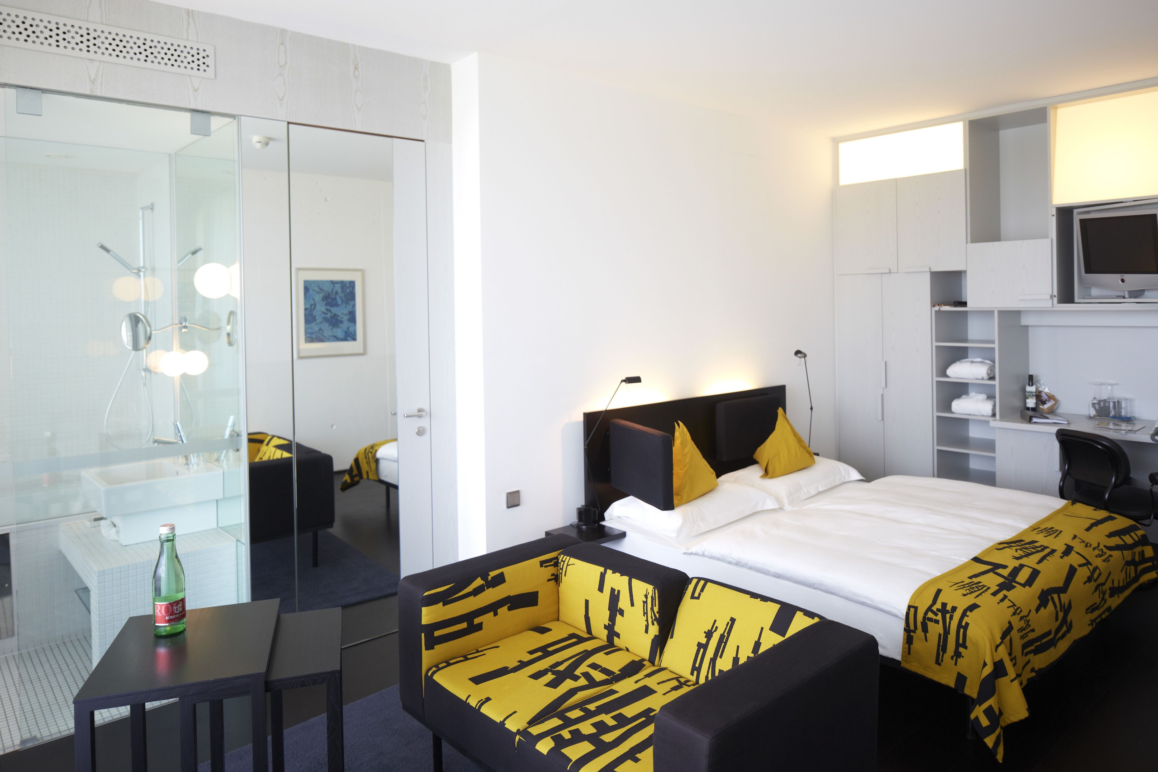 A modern hotel room with a glass shower enclosure, a bed with yellow and black patterned bedding, a small table, and a television on a shelf.