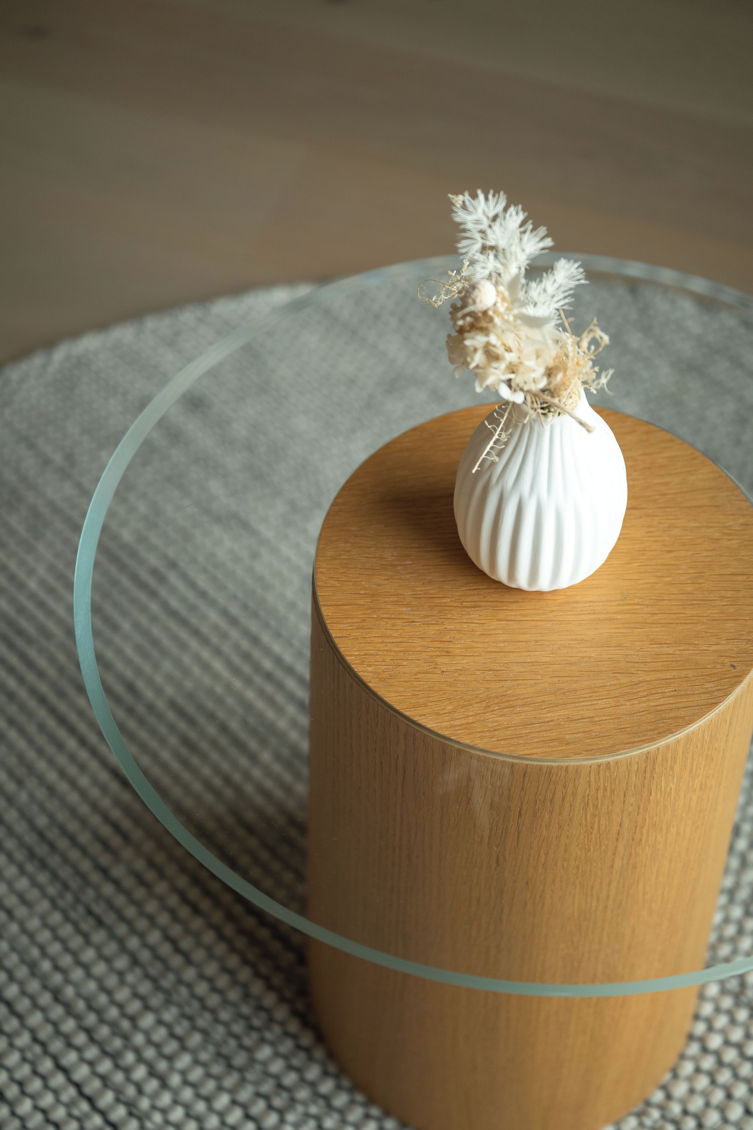 A round wooden table with a glass top, a white vase with dried flowers, and a textured grey rug.