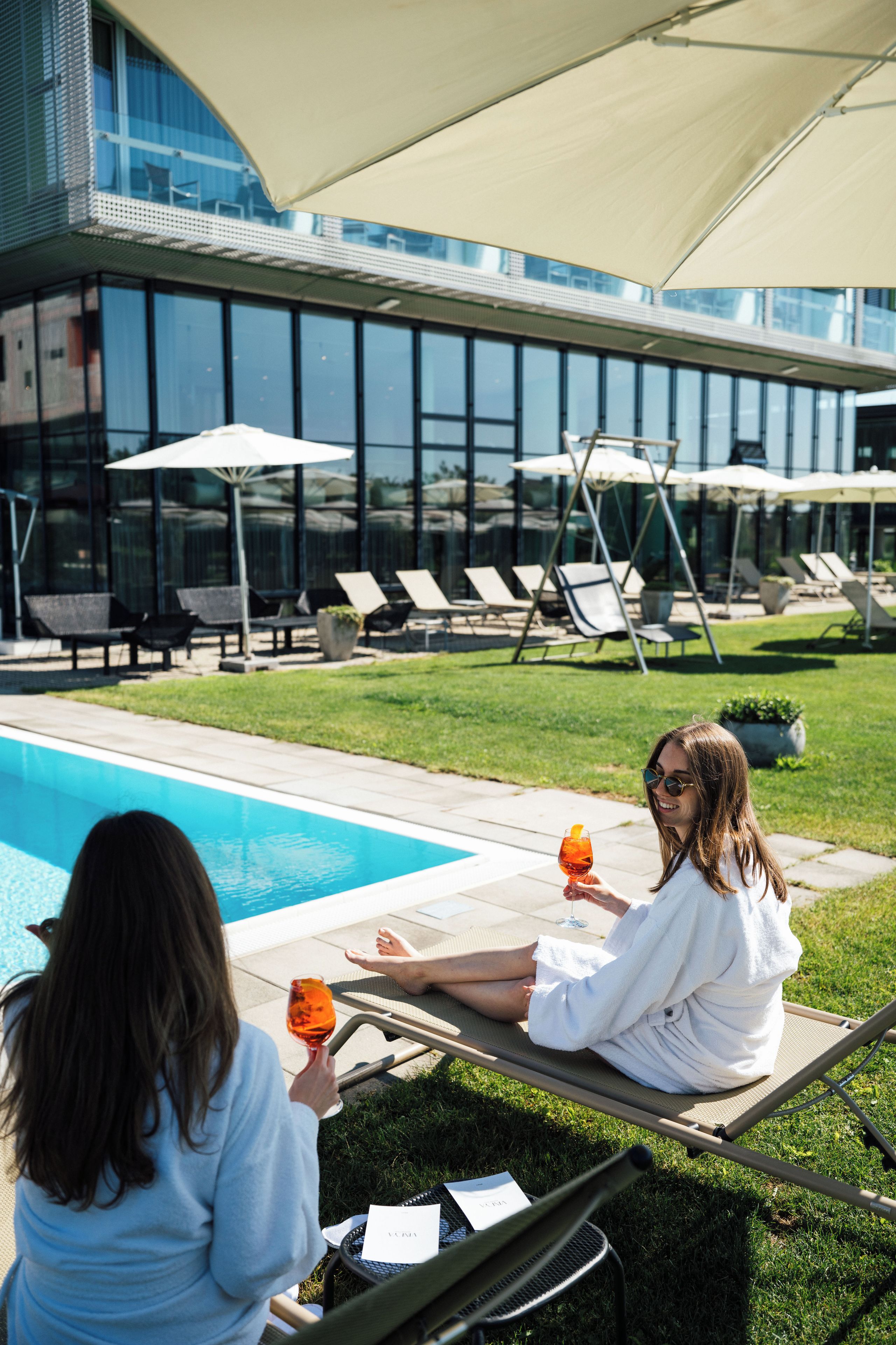Zona de piscina con sillones de descanso y sombrillas, una piscina azul cristalina y un edificio moderno con amplias ventanas al fondo.