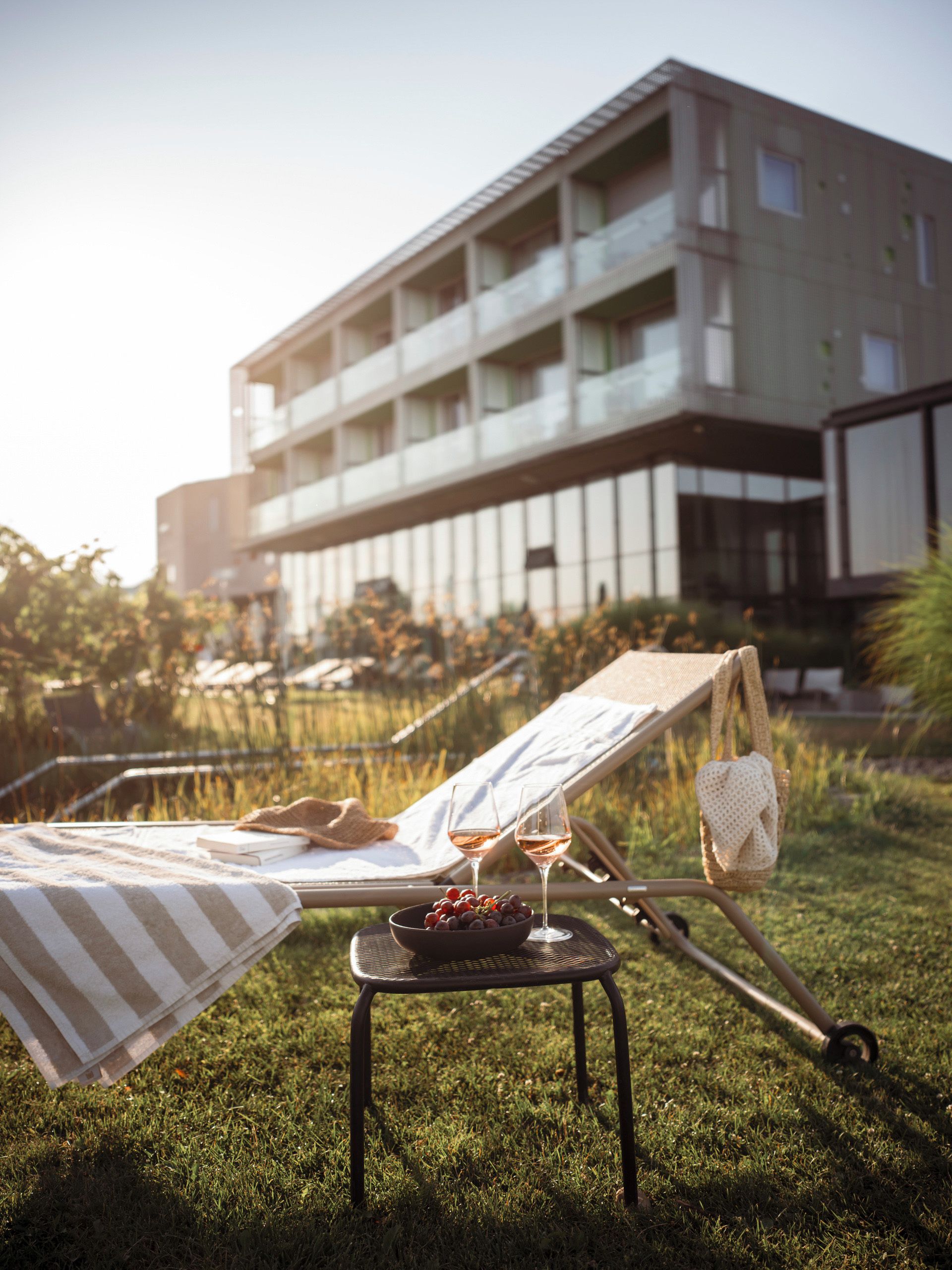 A lounge chair with a striped towel and a bowl of fruit, a wine glass, and a handbag on a small table in a grassy area with a building in the background.