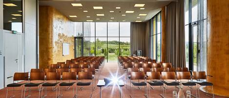 Un salón de conferencias con filas de sillas, una larga mesa y amplios ventanales que dan a vistas de zonas verdes.