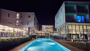 Une piscine entourée de chaises longues et de parasols, elle-même bordée d’immeubles modernes dotés de grandes fenêtres et de balcons.