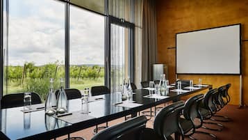 A conference room with a long table, chairs, a projector screen, and a view of greenery outside.
