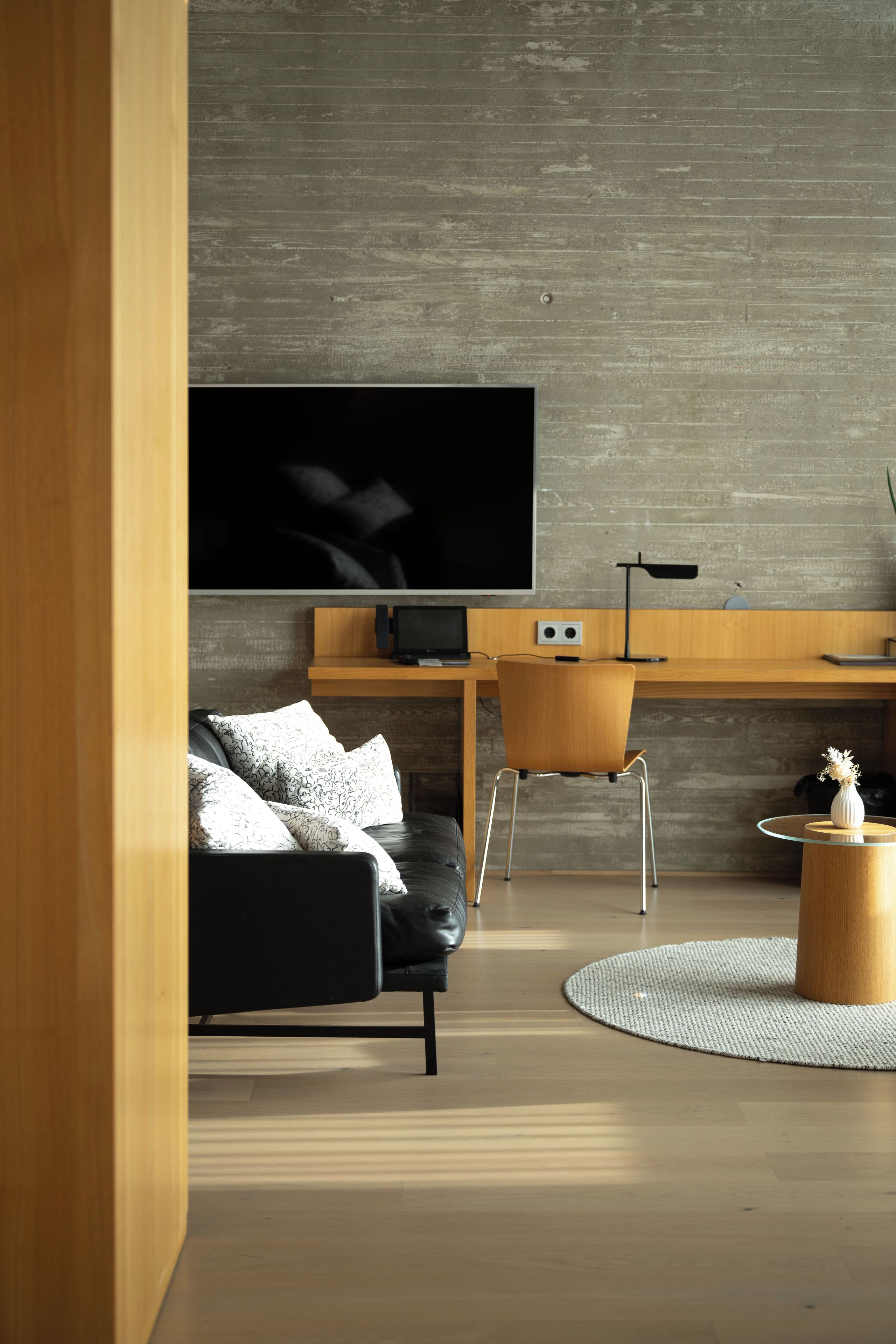 A modern living room with a flat-screen TV mounted on a concrete wall, a black leather sofa, a wooden desk with a computer, and a round wooden coffee table with a vase.