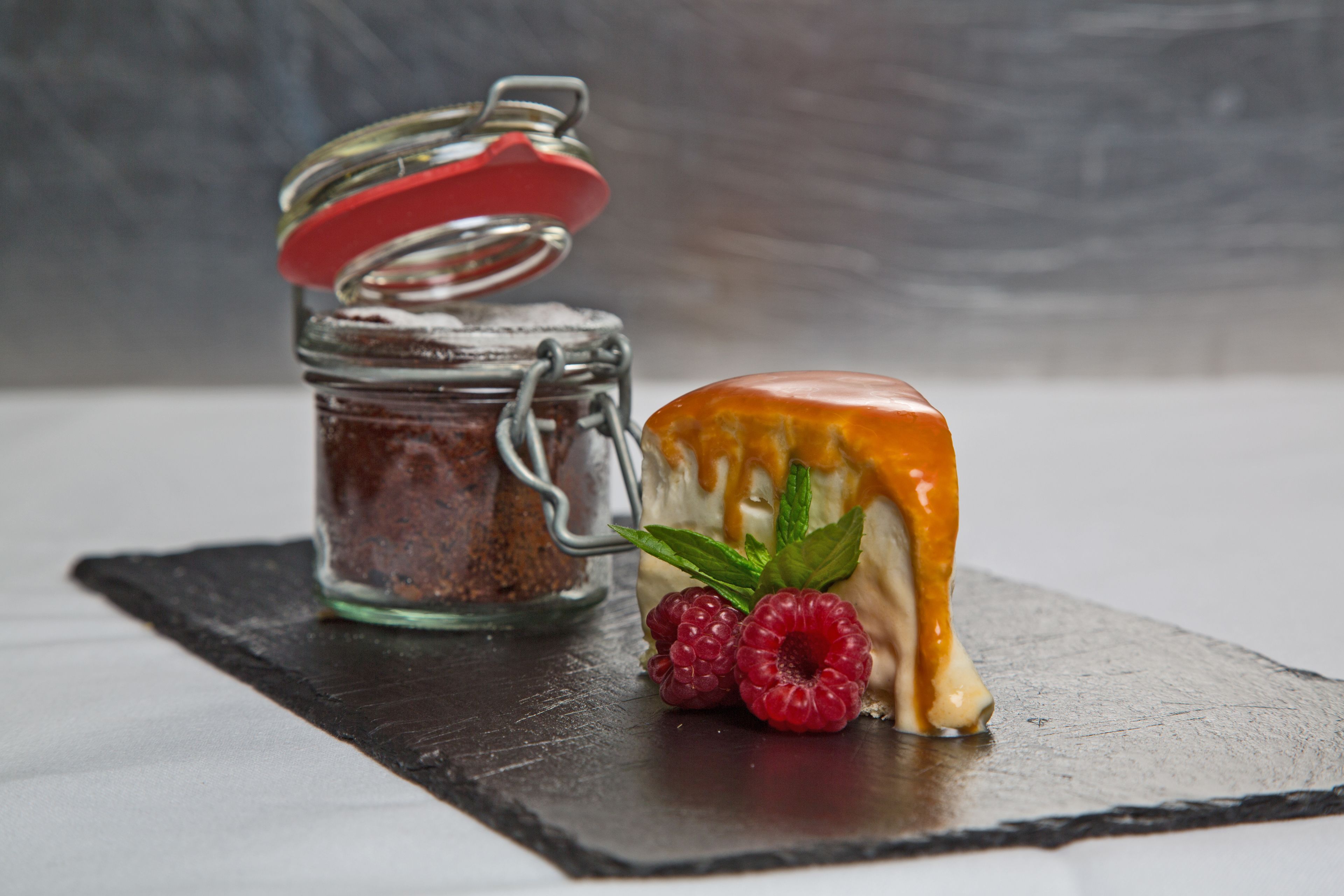 A dessert with a caramel drizzle and raspberries on a slate plate, next to a jar with a red lid.