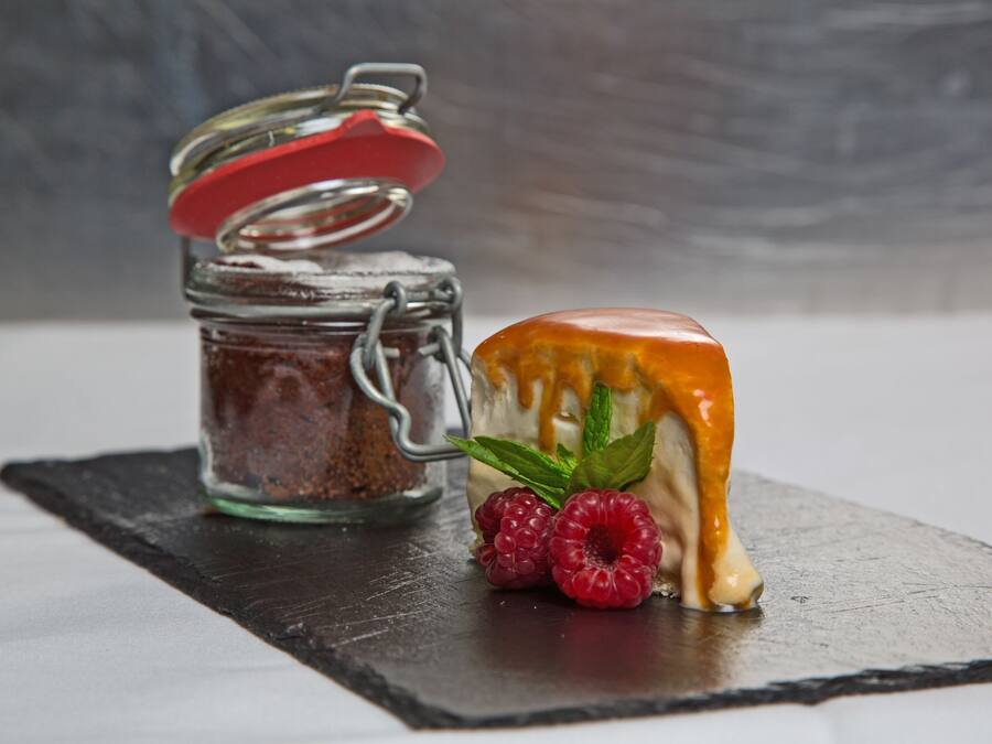 A dessert with a caramel drizzle and raspberries on a slate plate, next to a jar with a red lid.