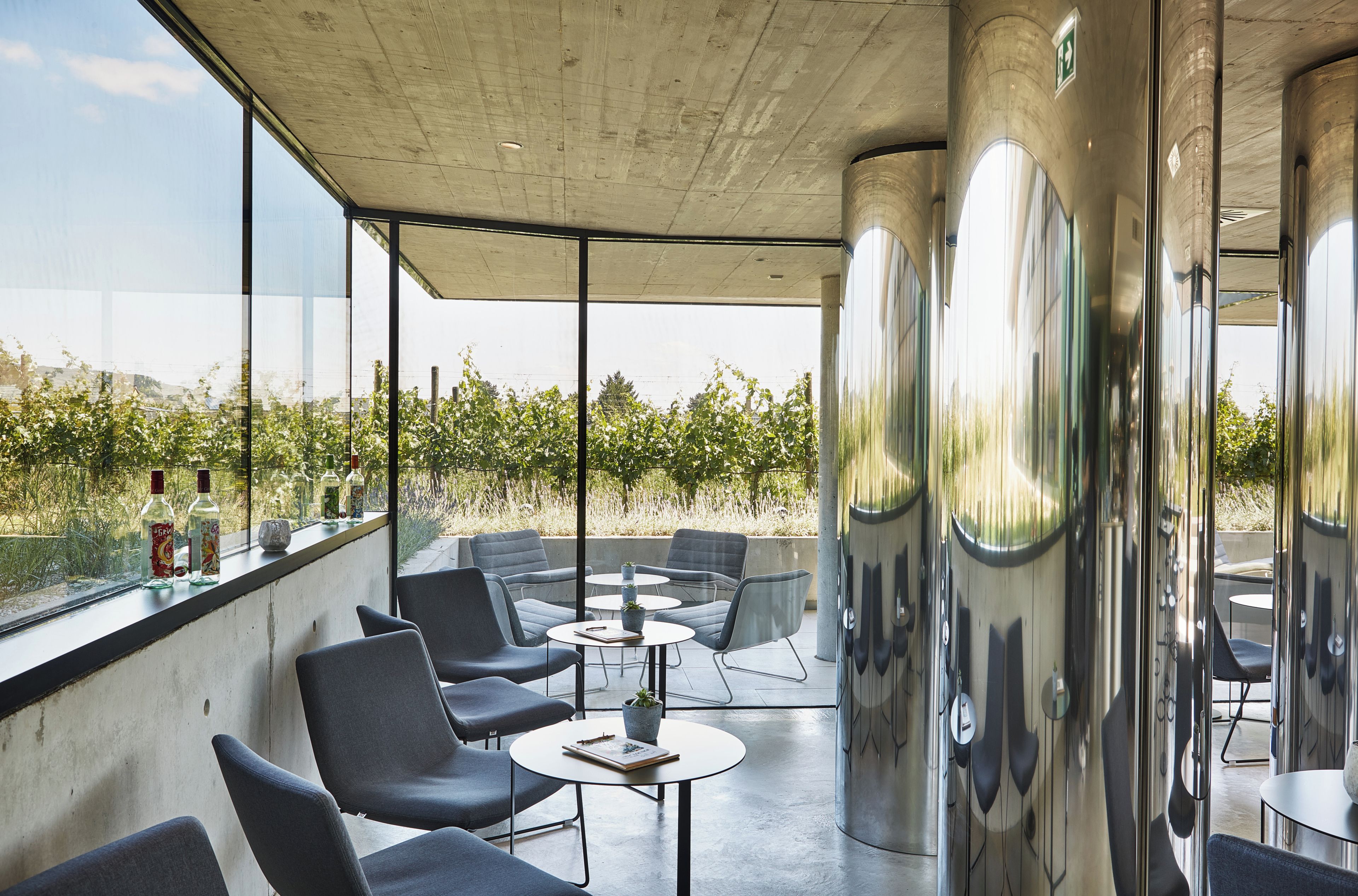 Un área de estar exterior moderna con sillas y mesas, ventanas amplias y vistas a un viñedo.