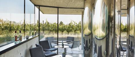 Un área de estar exterior moderna con sillas y mesas, ventanas amplias y vistas a un viñedo.
