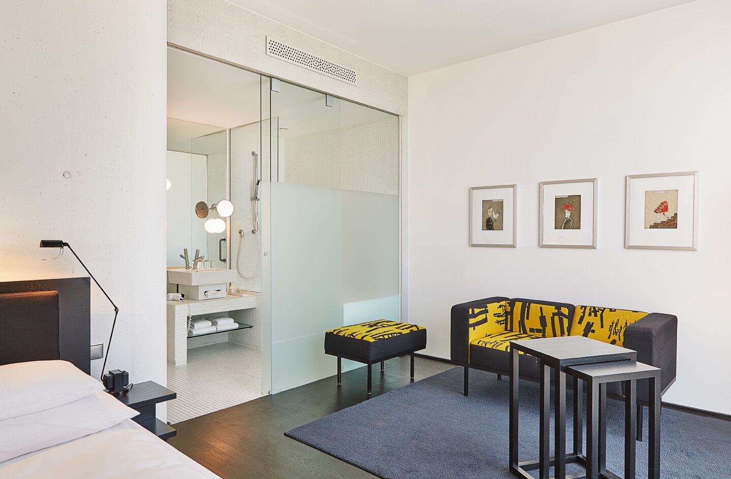 A modern hotel room with a bed, a desk, a sofa, and a shower area.