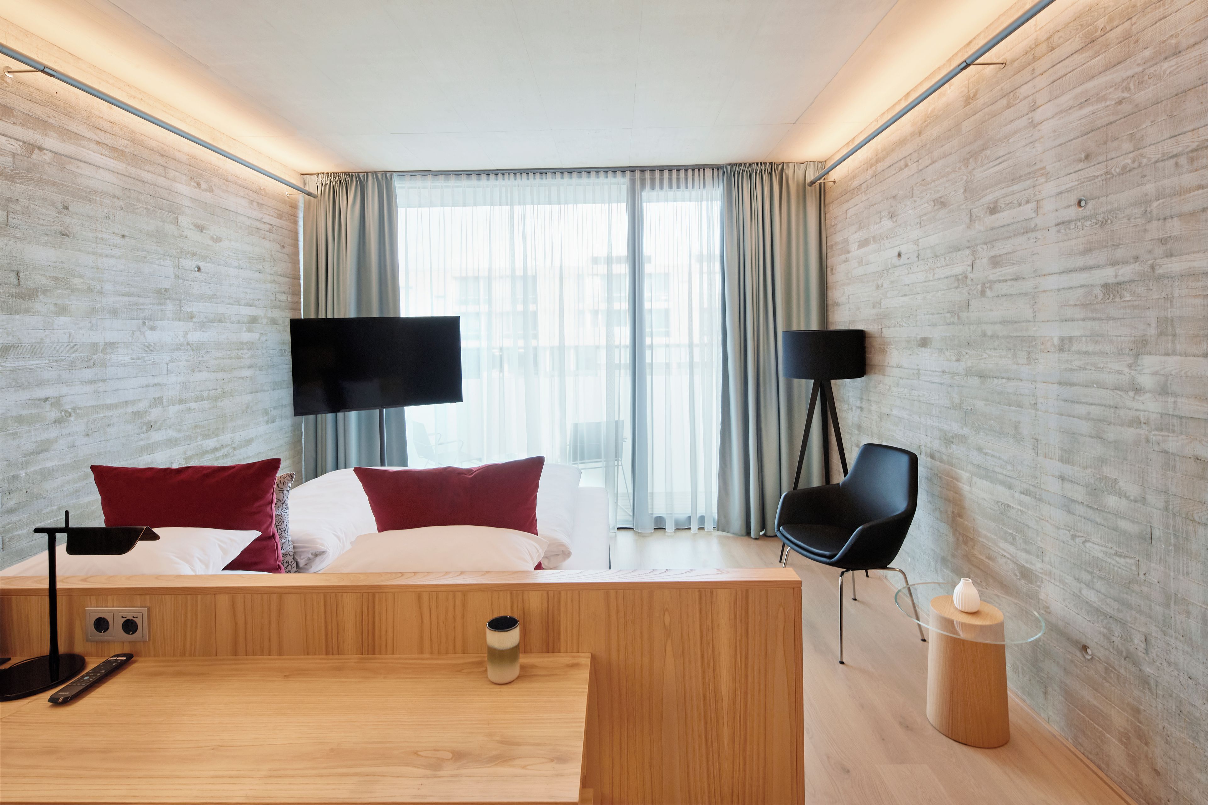 A modern hotel room with a wooden bed, a desk, a chair, and two black floor lamps.