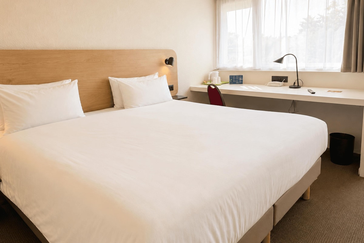 Een hotelkamer met een groot bed, een bureau, een stoel, een lamp en een raam met gordijnen.