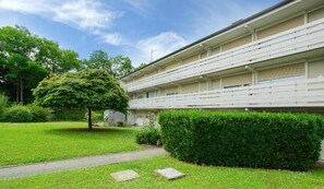 Ein mehrgeschossiges Gebäude mit flachem Dach, einem gepflasterten Weg und einem gepflegten Rasen mit Baum und Hecke.
