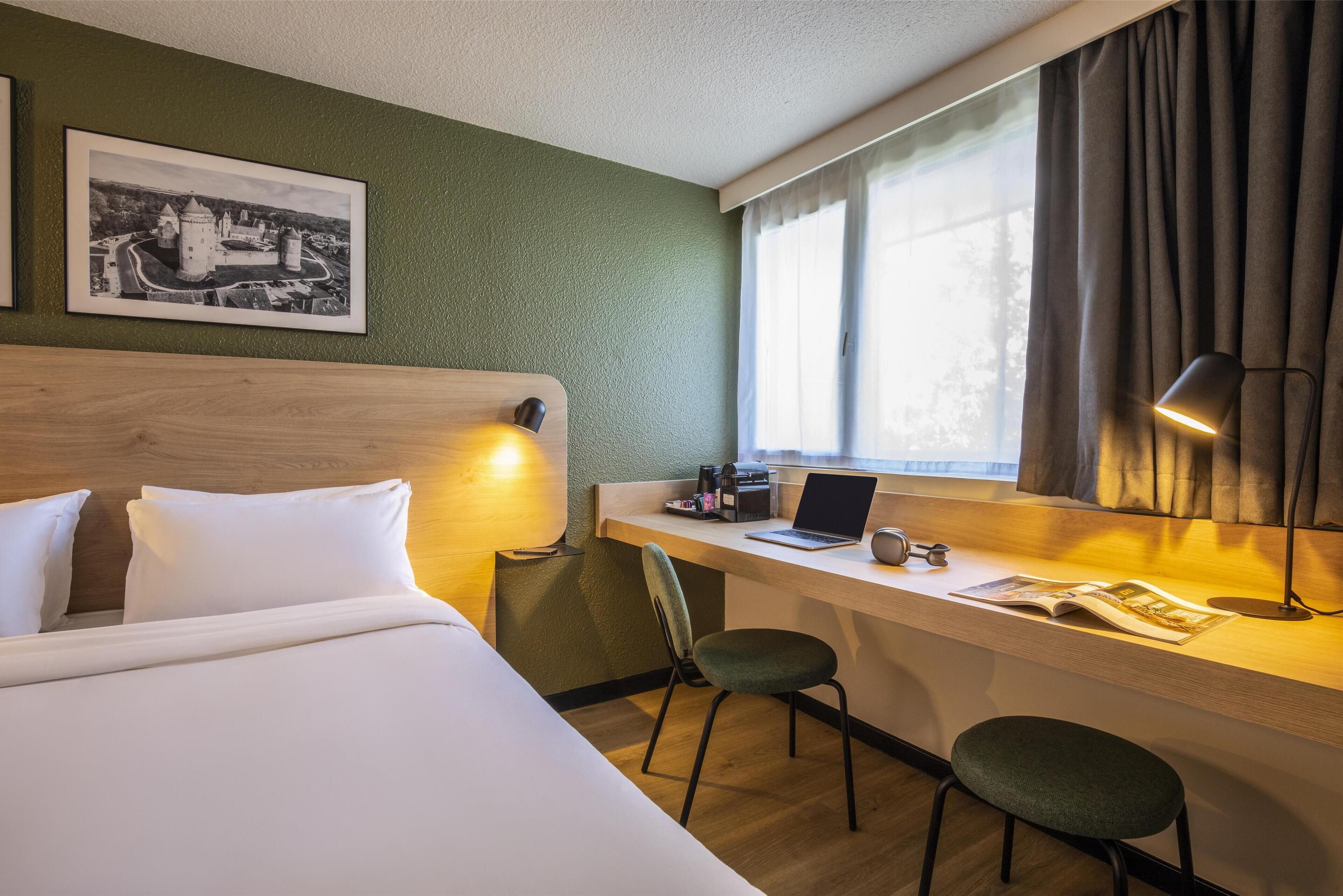 Une chambre d’hôtel avec un lit, un bureau avec un ordinateur portable, une chaise, une lampe et une fenêtre avec des rideaux.