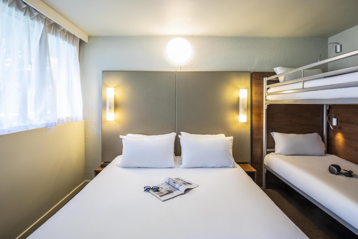 Quarto de hotel com uma cama grande, duas mesas de cabeceira, uma cama beliche e uma janela com cortinas.