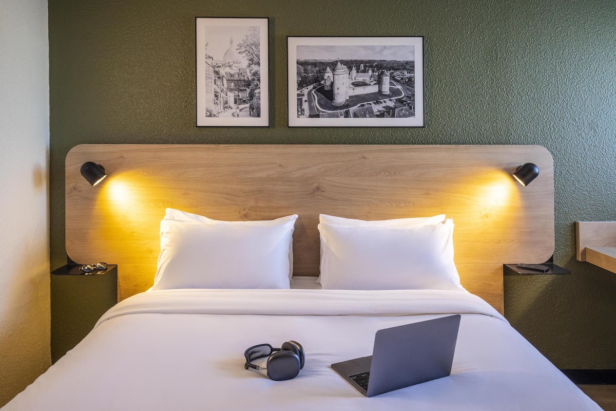 Un lit bien fait, avec des draps blancs, un ordinateur portable et des écouteurs posés dessus. Deux tableaux encadrés sont accrochés au mur, au-dessus du lit.