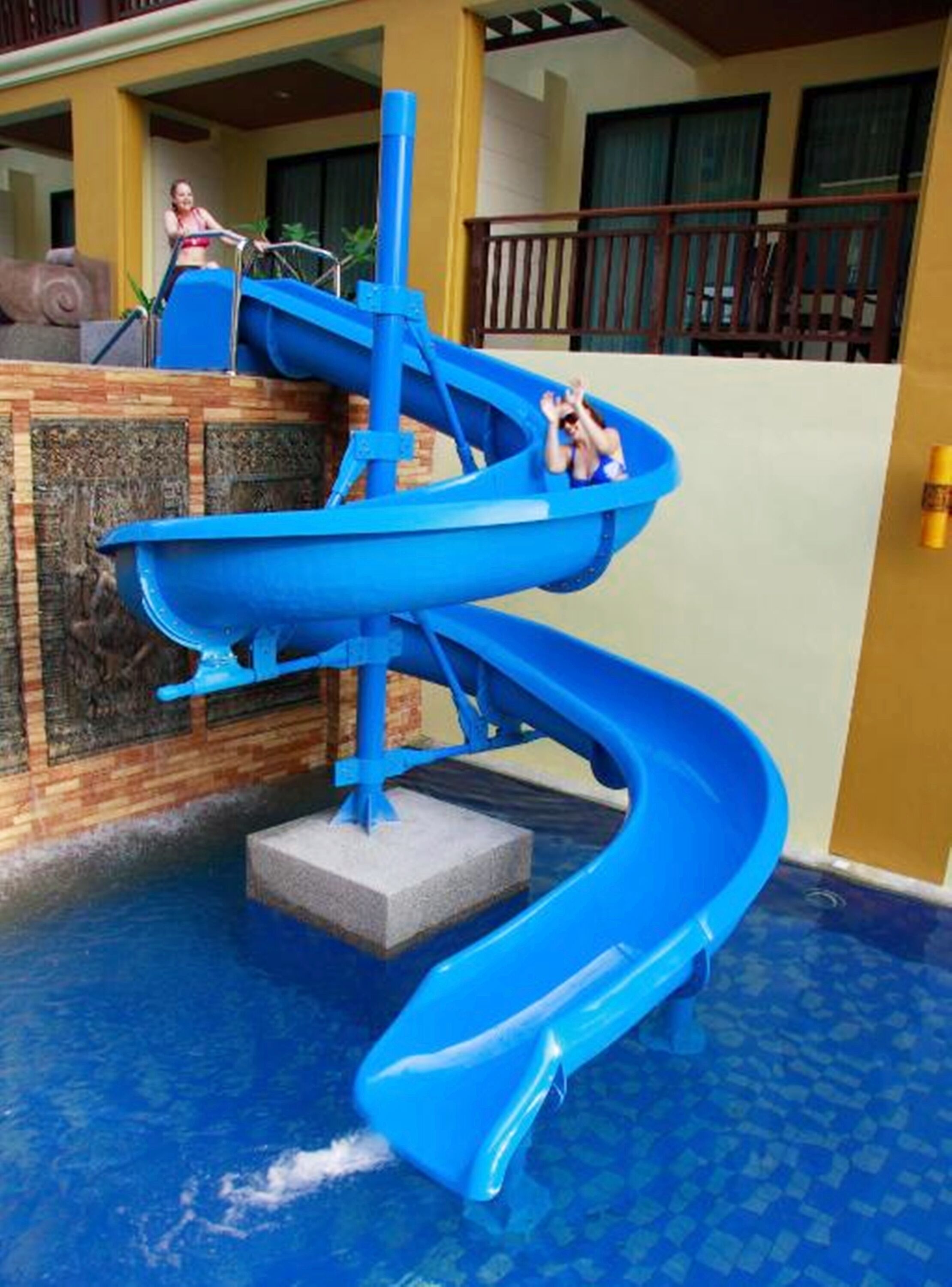 A blue water slide in a pool area with a wooden deck and a yellow building in the background.