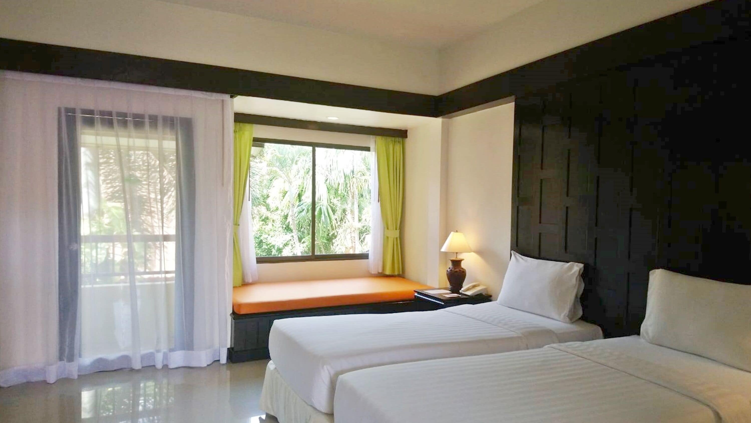 Habitación de hotel con dos camas, una ventana con cortinas, una mesita de noche con lámpara y vistas al verdor exterior.