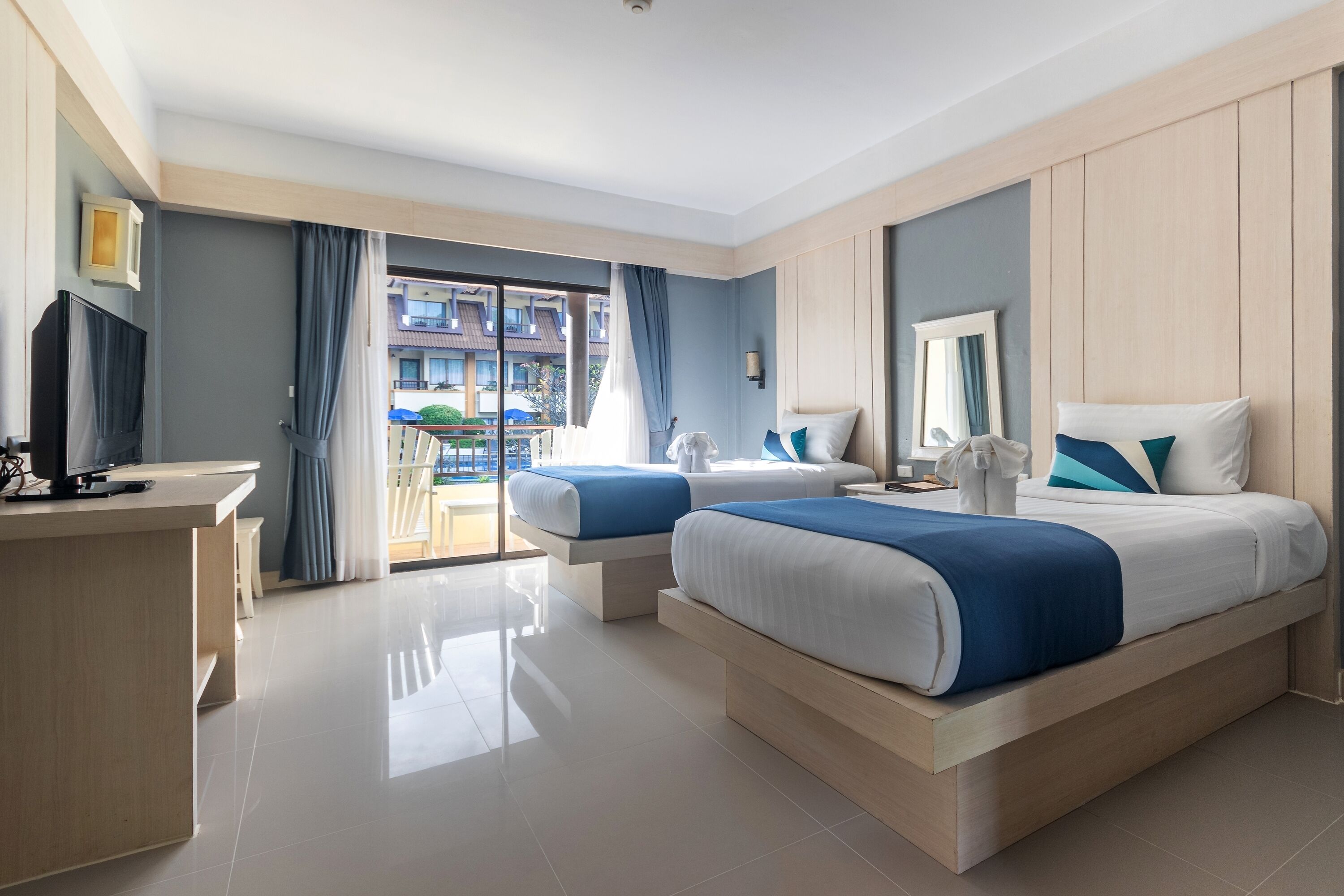 A hotel room with two beds, a desk, a TV, and a balcony with a view of buildings.