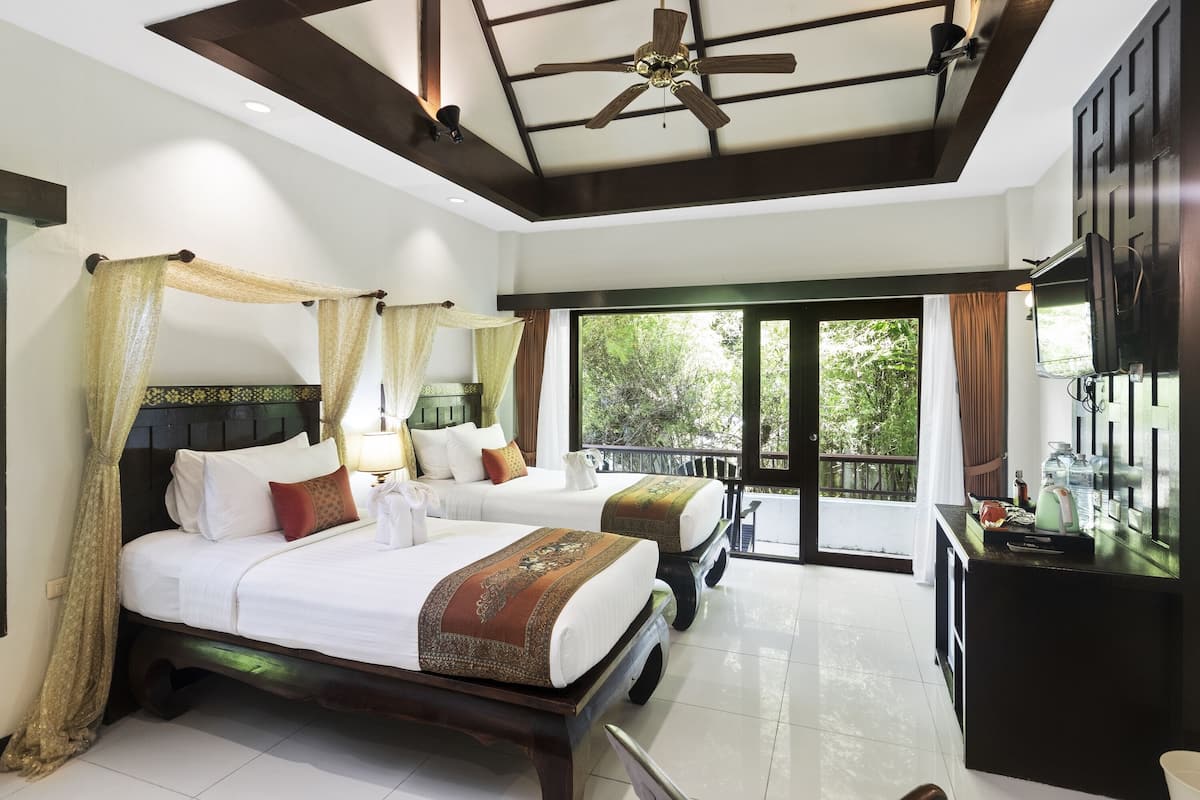 A hotel room with two beds, a ceiling fan, a TV, and a balcony with a view of greenery.