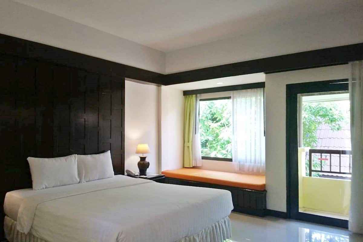 A hotel room with a large bed, two bedside lamps, a window with a view of greenery, and a balcony with a bench.