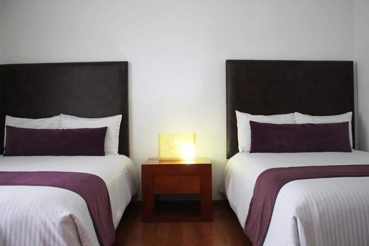 Habitación de hotel con dos camas, cada una con cabecera, mesita de noche con lámpara y una manta morada.