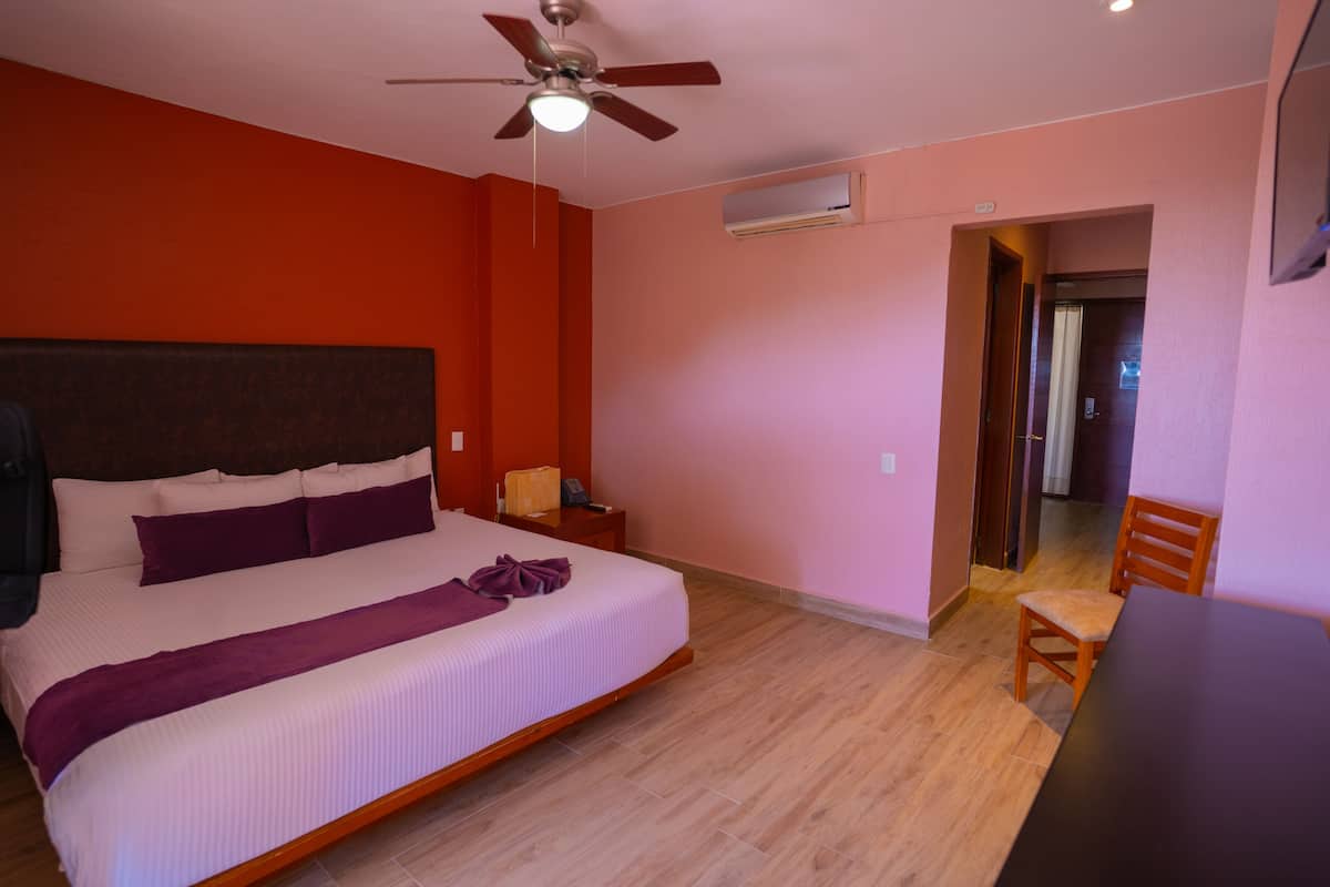 Un dormitorio con cama, ventilador de techo, unidad de aire acondicionado, mesita de noche de madera, una silla y una puerta que da a otra habitación.