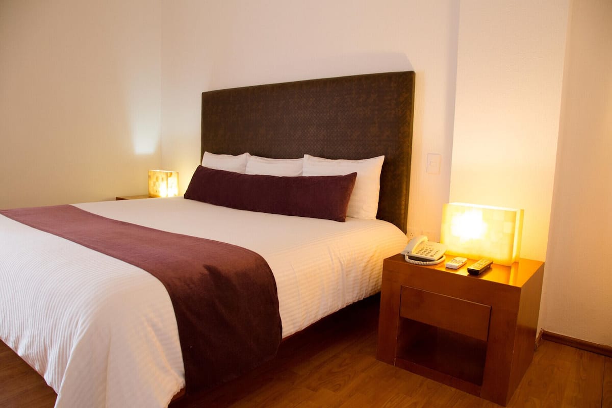Una cama bien tendida con cabecera, lámparas de noche y una mesita de noche con teléfono y controles remotos.