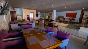 Un'area pranzo moderna con sedute viola, tavoli in legno e una cucina sullo sfondo.