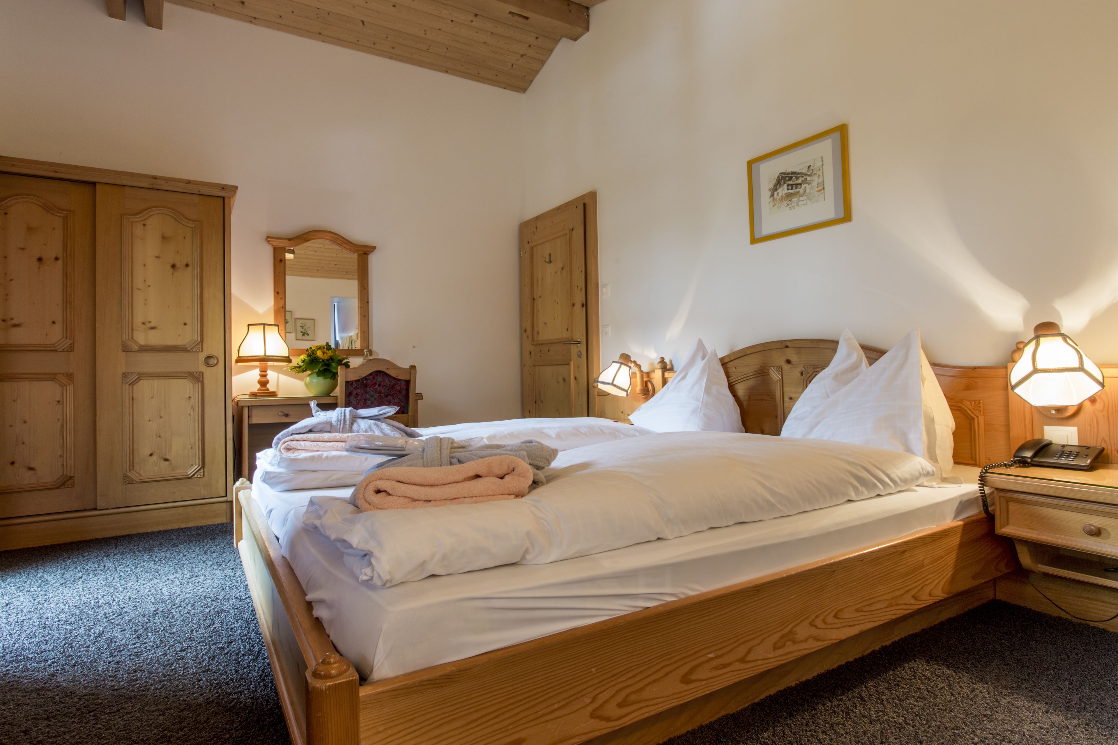 A neatly made bed with white linens and a pink towel, wooden headboard, bedside lamps, a wooden nightstand, and a wooden wardrobe.