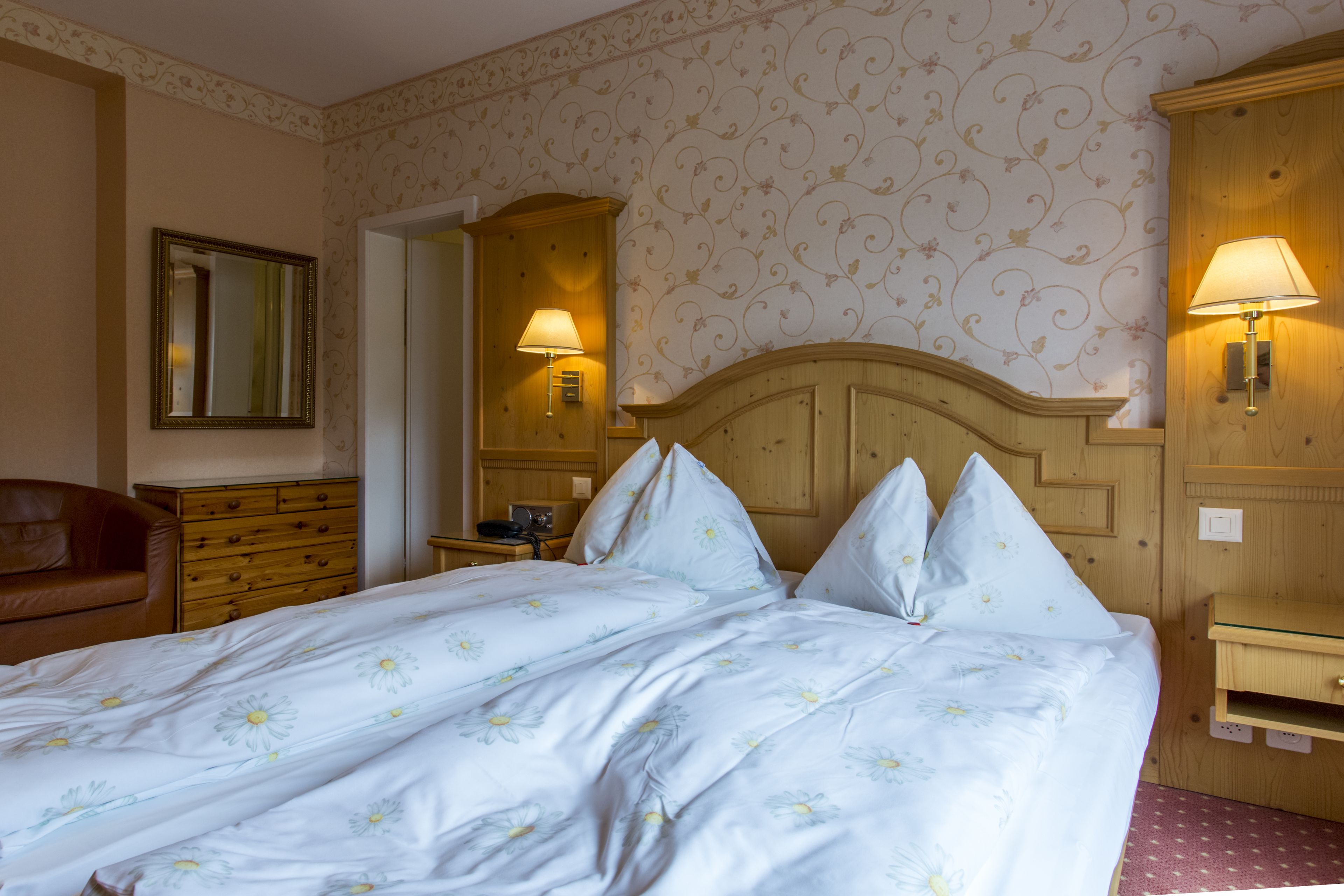 Habitación de hotel con una cama grande, cabecera de madera, papel tapiz floral, dos lámparas de noche, una mesita de noche, un aparador y un espejo.