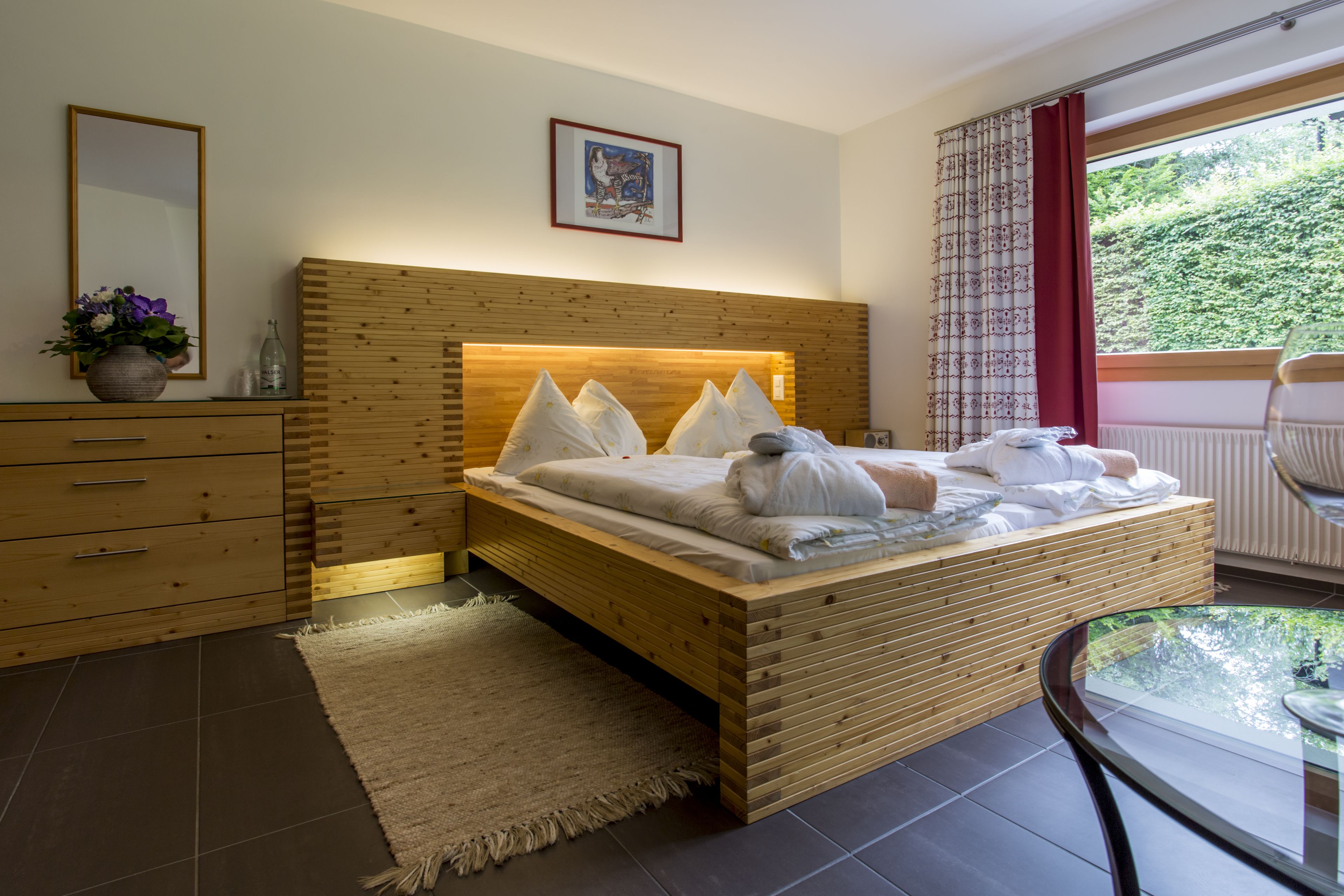 A modern bedroom with a wooden bed, a bedside table with a vase of flowers, a mirror, and a window with a view of greenery.