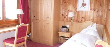 A wooden room with a bed, a desk, a chair, a wardrobe, and a nightstand with a lamp.