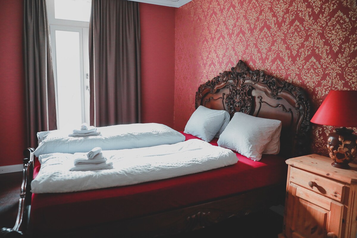 Ein ordentlich gemachtes Bett mit weißen Leinen und Kissen, ein Nachttisch aus Holz mit einer Lampe und eine Wand mit rotem Muster.