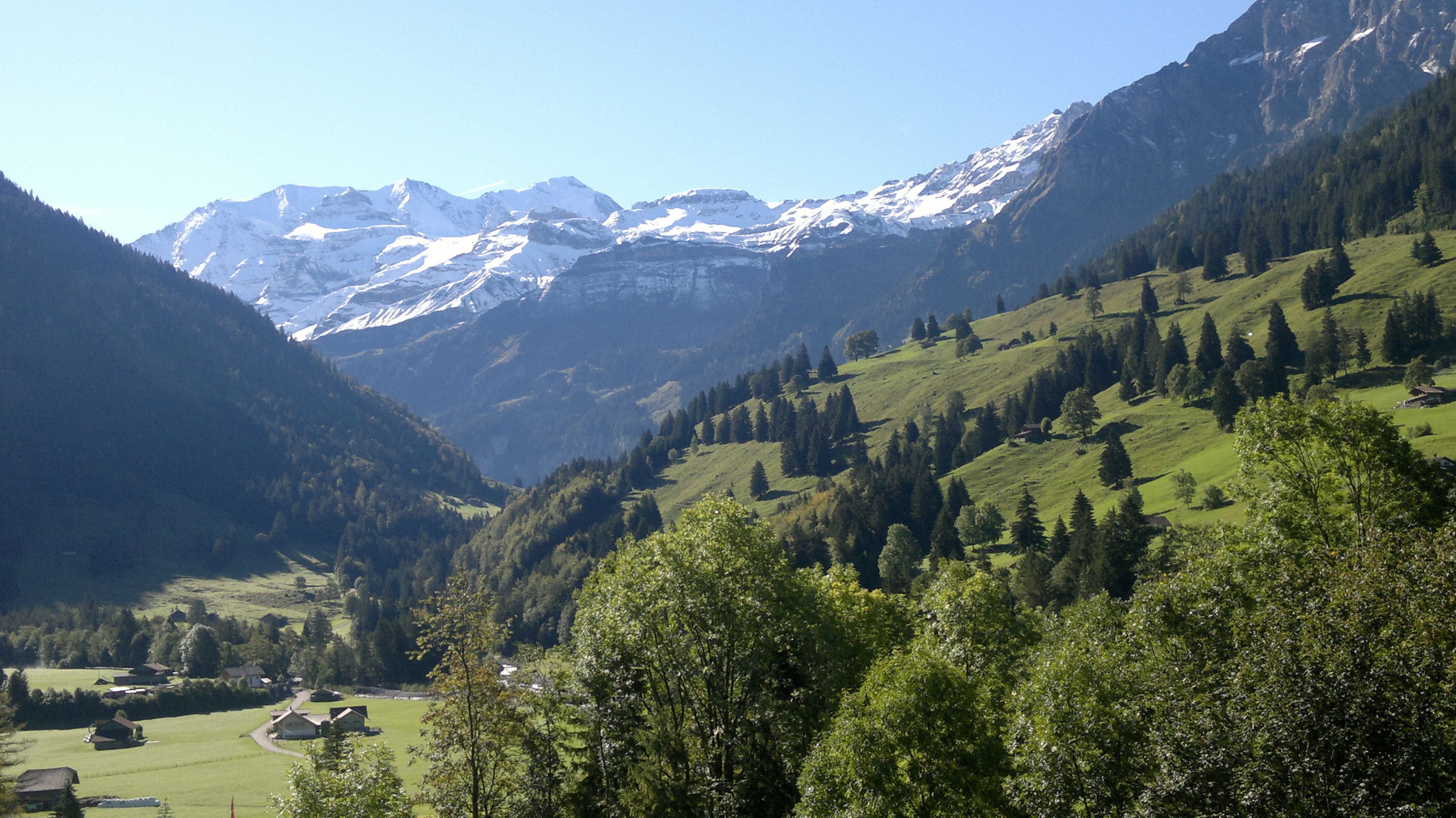 A mountain landscape with snow-capped peaks, a valley with green fields, and scattered houses.