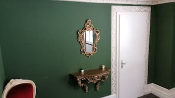 A room with green walls, a white door, a red chair, a gold-framed mirror, and a small table with a plant.