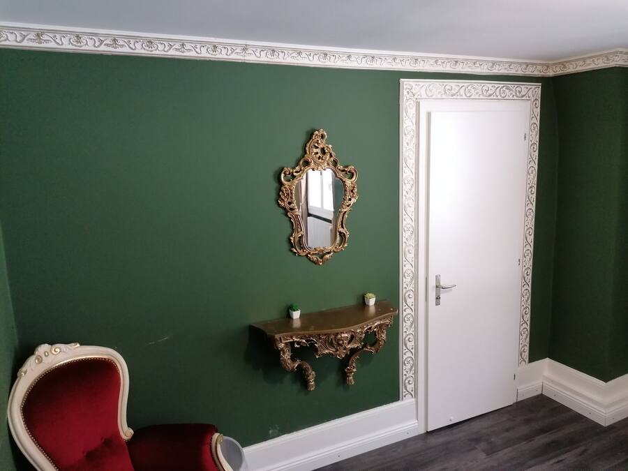 A room with green walls, a white door, a red chair, a gold-framed mirror, and a small table with a plant.
