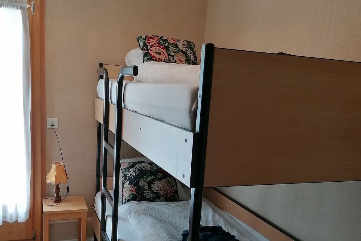 A bunk bed room with a small table and lamp, wooden flooring, and a window with curtains.