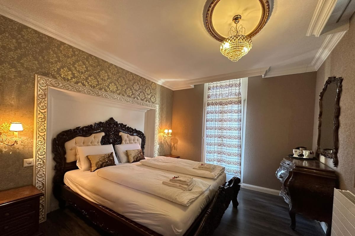 A classic hotel room with a large bed, ornate headboard, and a chandelier.