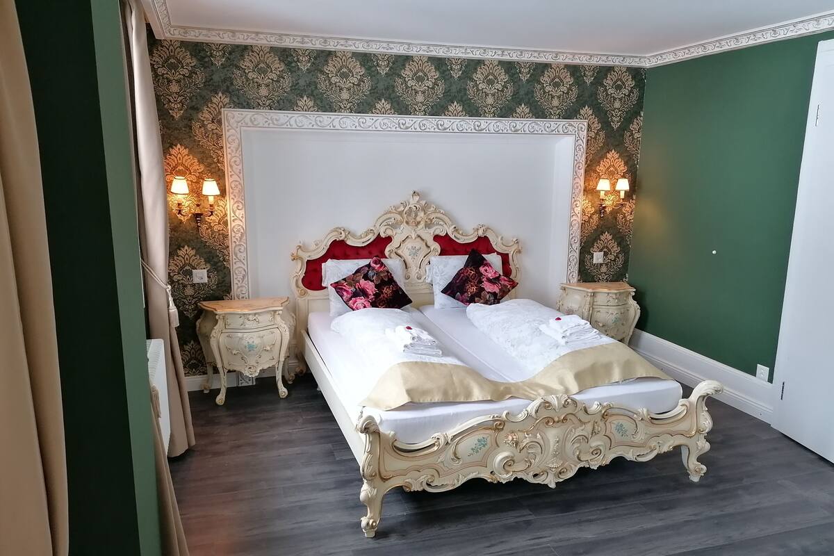 A classic four-poster bed with ornate headboard and footboard, flanked by two nightstands with lamps, in a room with patterned wallpaper and dark wooden flooring.