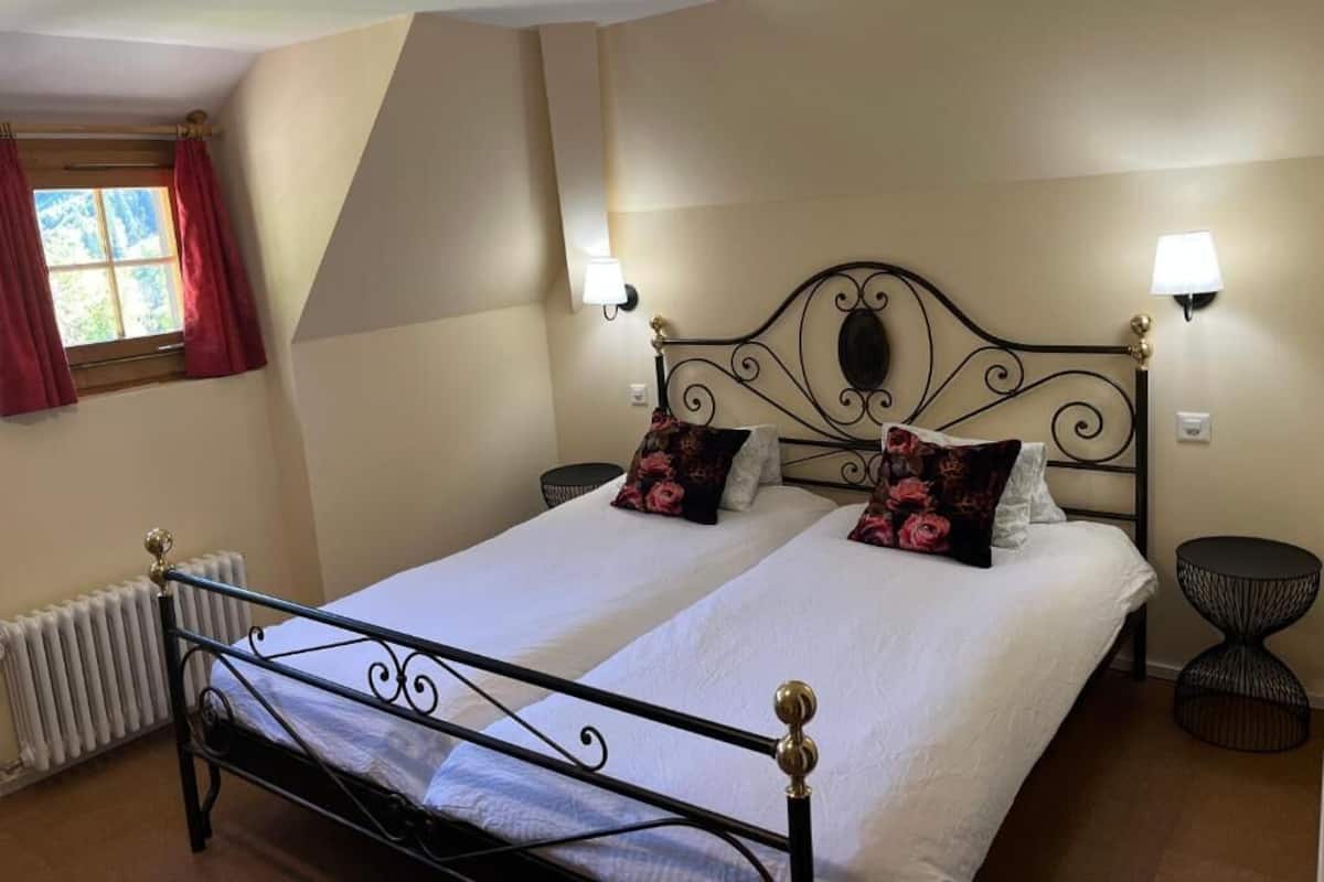 A double bed with a metal frame, two wall-mounted lamps, a radiator, a window with curtains, and a round table.