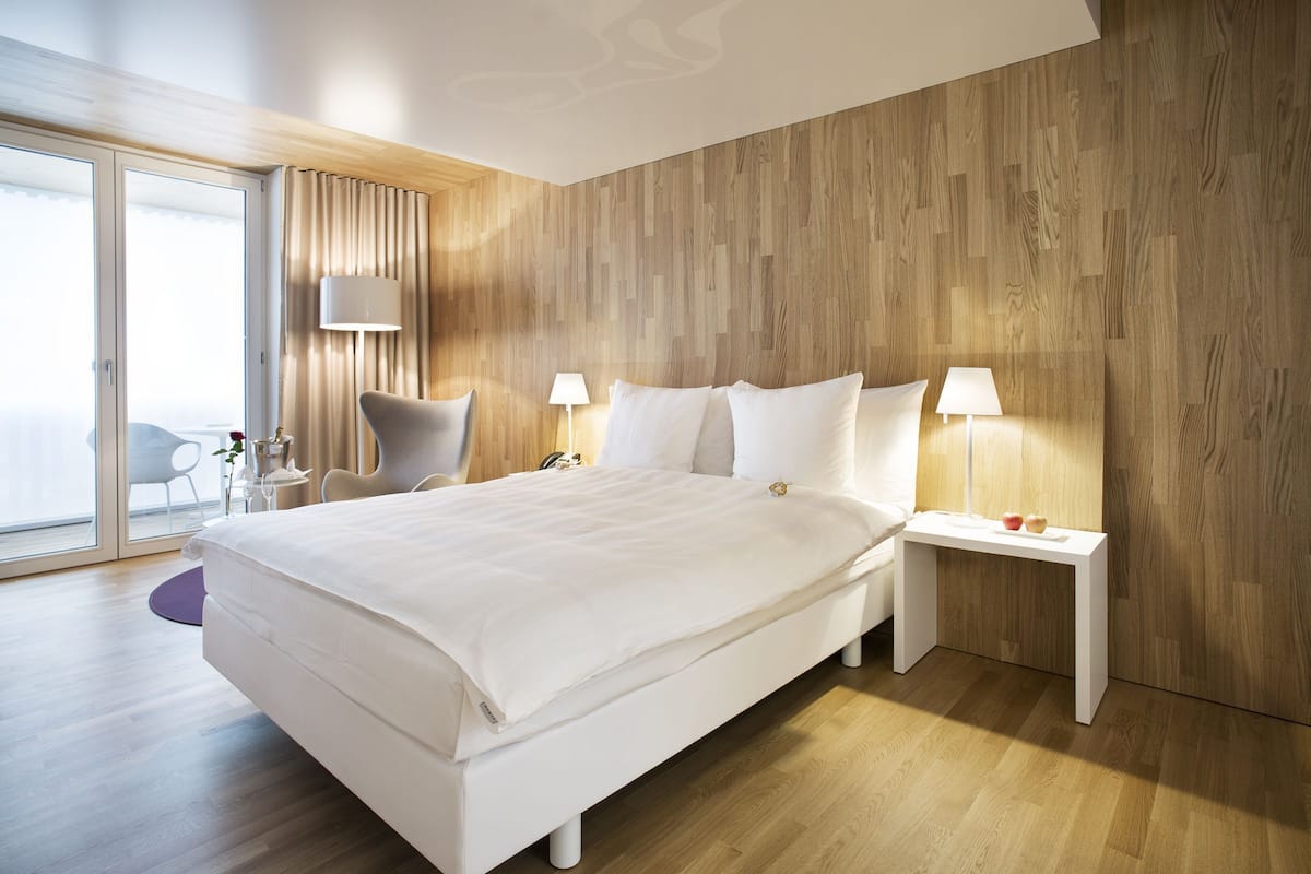 A modern hotel room with a large bed, two bedside tables with lamps, a chair, and a view through a sliding glass door.