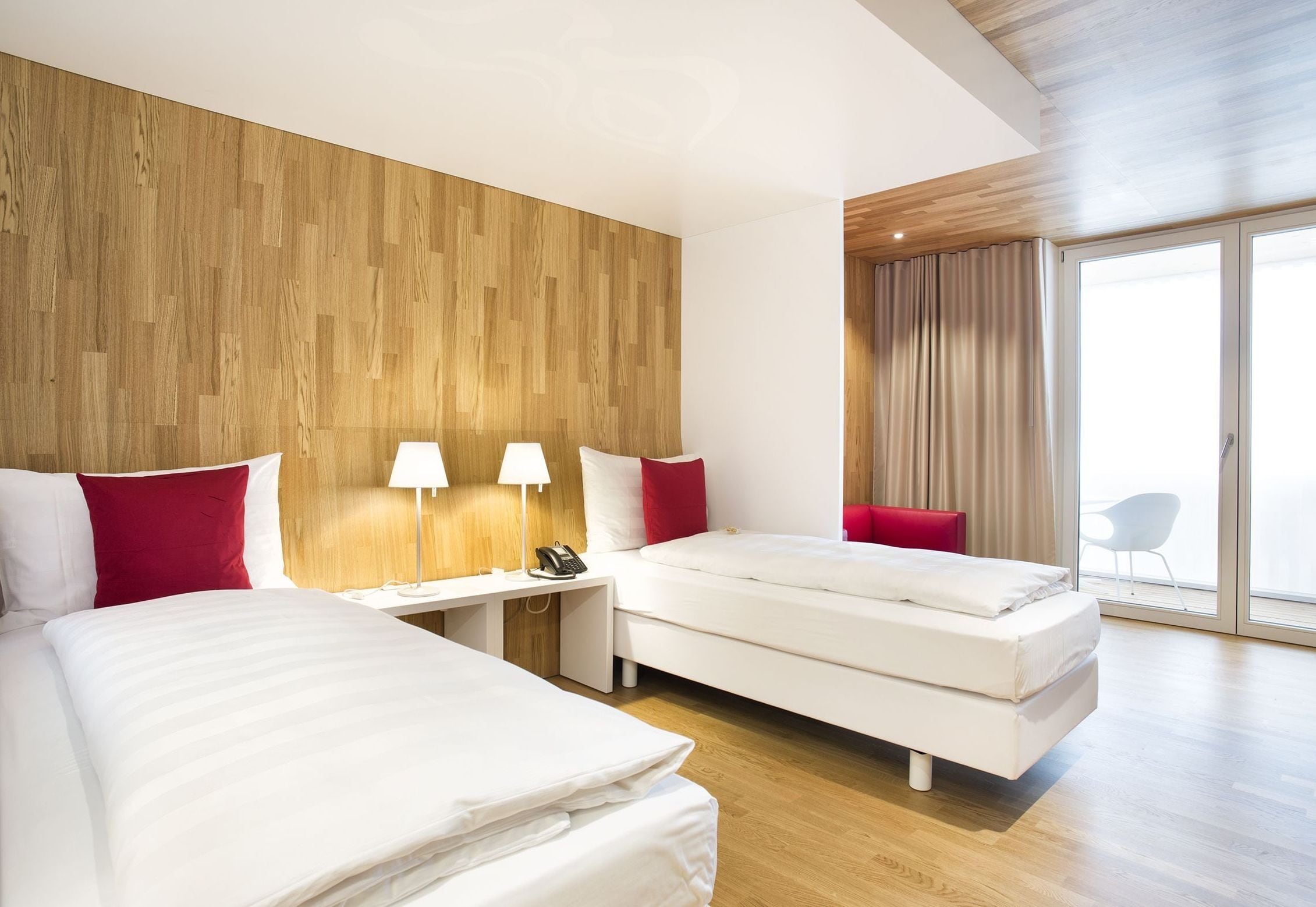 A modern hotel room with two beds, a wooden accent wall, a desk with a lamp, and a large window with curtains.