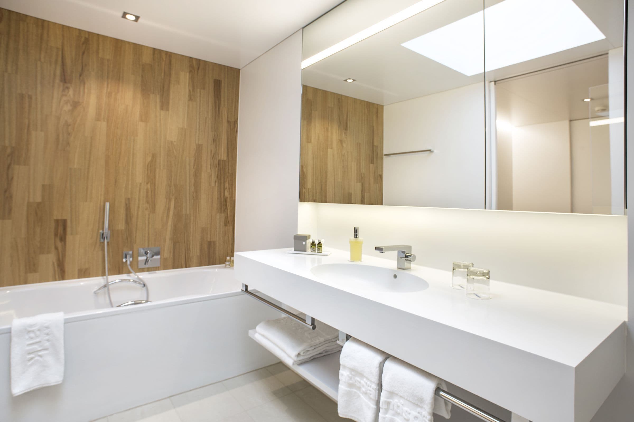 A modern bathroom with a wooden wall, a large mirror, a white sink, and a bathtub.