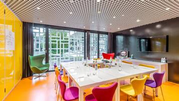 Un salón de conferencias con una mesa blanca, sillas coloridas, un diseño moderno de techo y grandes ventanas con vistas a zonas verdes.