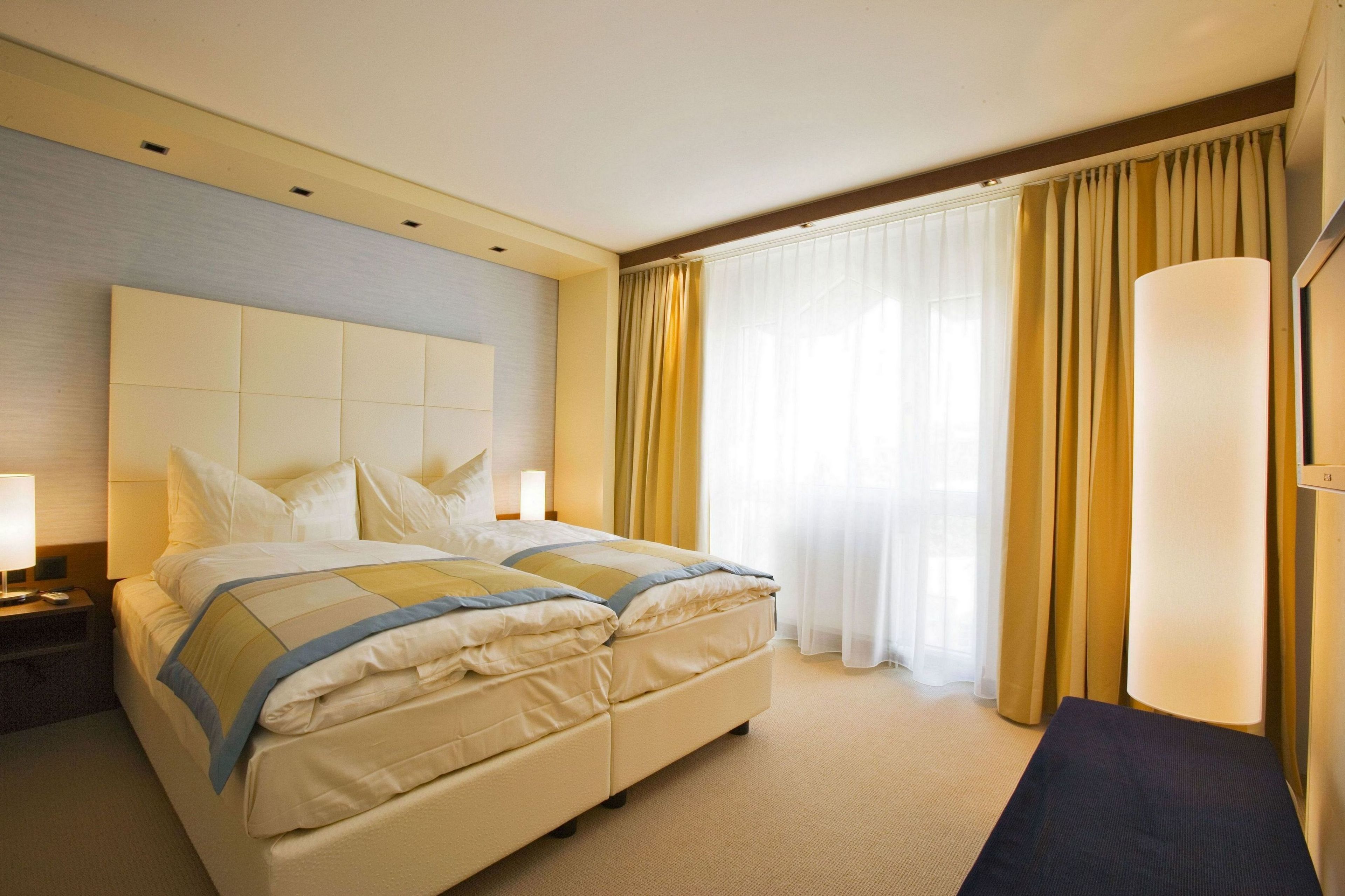 A hotel room with a large bed, two bedside tables, a desk, and a large window with curtains.