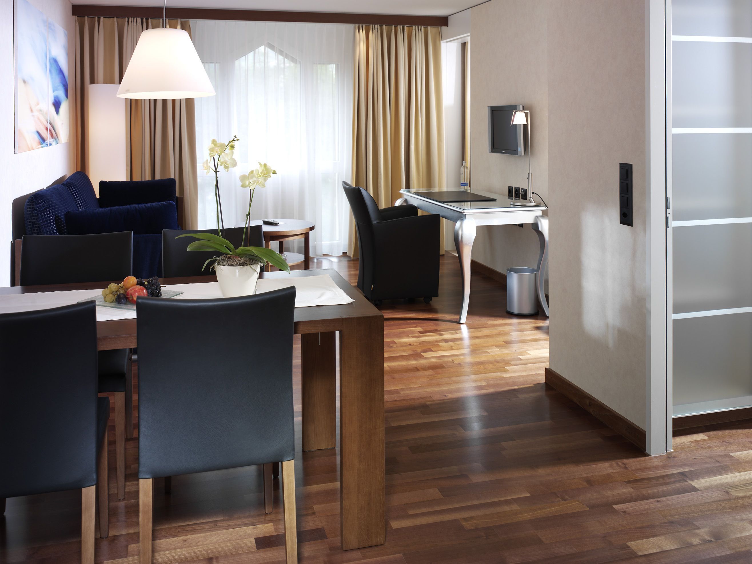 A modern hotel room with a dining area, a sofa, a TV, and a plant.