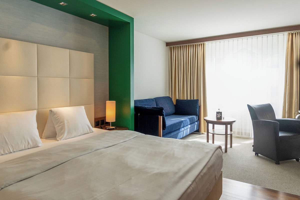 A hotel room with a large bed, a sofa, a chair, a small table, and a lamp.