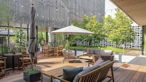 Un patio exterior con mobiliario de madera, un paraguas grande y una estatua.